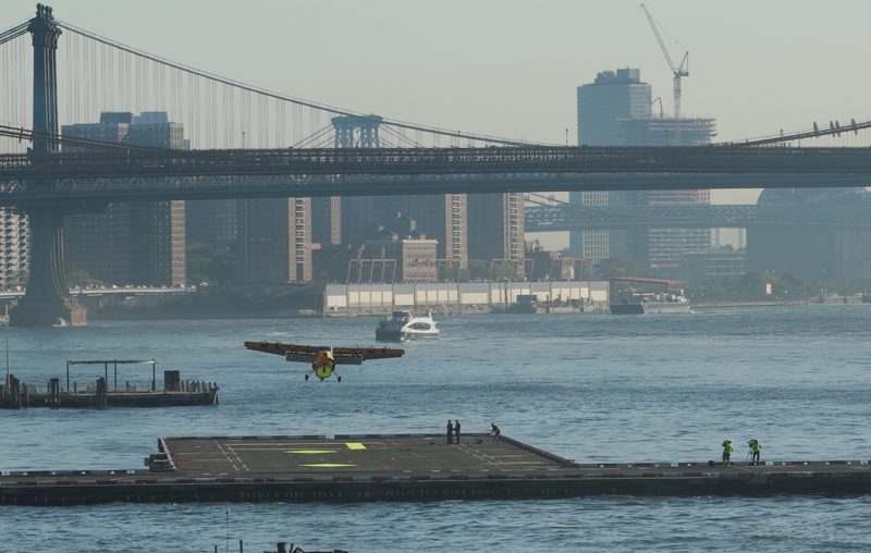 Electra's small hybrid-electric airplane flying low over the East River toward the Downtown Manhattan Skyport helipad, with the Brooklyn Bridge behind
