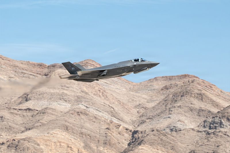 An F-35A fighter jet climbing past a desert ridge